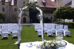 Historické nádvoří zámku Libouchec připravené pro letní svatbu s bílými židlemi a květinovou bránou.