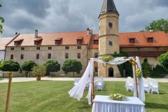 Historické nádvoří zámku Libouchec připravené pro letní svatbu s bílými židlemi a květinovou bránou.